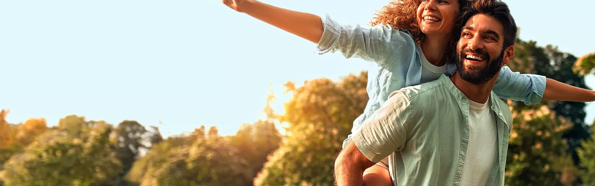 Ein Pärchen genießt einen sonnigen Tag im Park. Der Mann trägt seine Frau auf dem Rücken und sie hat lachend die Arme weit ausgebreitet.