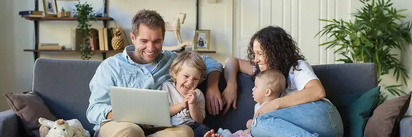 Eine Familie mit zwei kleinen Kindern sitzt zusammen auf einem Sofa. Alle lachen. Der Mann hat einen Laptop auf dem Schoß und scheint seiner Familie dort etwas zu zeigen.