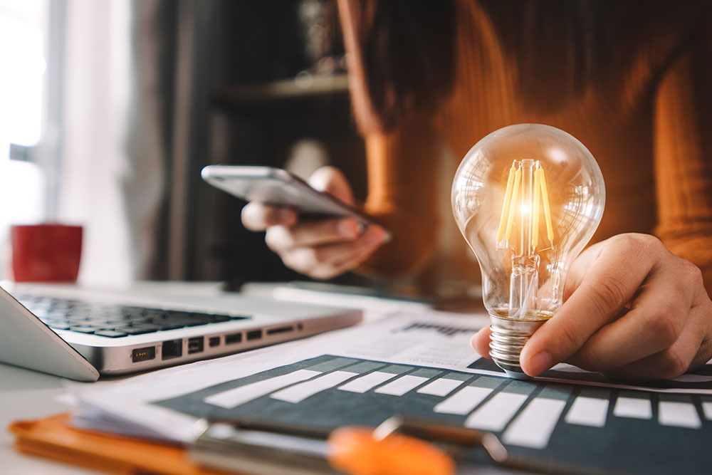 Leuchtende Glühbirne in der Hand einer Person mit Handy und Laptop symbolisiert Partnerschaft Energieversorgung